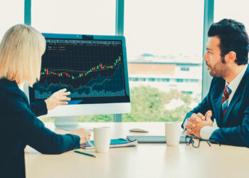 Two business professionals sit at a desk discussing a computer screen displaying a financial chart with rising data lines. Both are dressed in formal attire and appear engaged in conversation. | FintechZoom