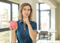 A woman in a denim dress holds a pink piggy bank and looks thoughtful, standing in a bright room with large windows and modern decor. | FintechZoom