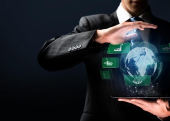 A man in a business suit holds a tablet with a glowing digital globe above it, surrounded by green industry and sustainability icons on a dark background. | FintechZoom