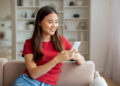 A young woman with long dark hair, wearing a red t-shirt and jeans, sits on a beige sofa and smiles while looking at her smartphone. A bright living room with shelves is visible in the background. | FintechZoom