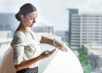 A woman in professional attire stands by a window in a modern office, looking at her smartphone. City buildings are visible outside the bright window in the background. | FintechZoom