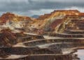 Terraced layers of an open pit mine with reddish and brown soil under a cloudy, overcast sky. The landscape shows exposed earth and rock, with puddles of water on some lower levels. | FintechZoom