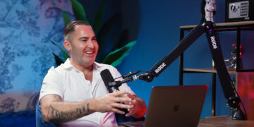 A man with tattoos on his left arm sits at a desk, smiling and speaking into a microphone on a boom arm. He faces a laptop, with a blue background, plants, and shelves behind him. | FintechZoom