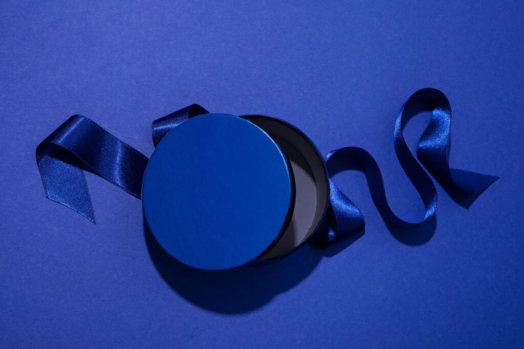 A round blue gift box with its lid slightly open, placed on a blue surface with a shiny blue ribbon extending from the box and curling to the side. | FintechZoom