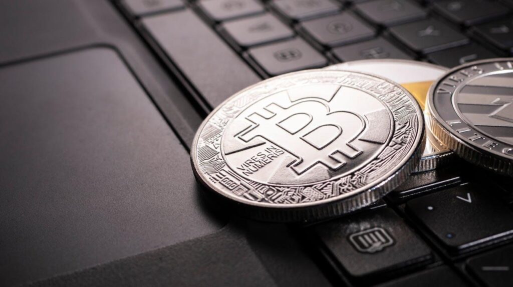 A close-up of physical Bitcoin and Litecoin coins resting on a laptop keyboard, symbolizing digital currency and cryptocurrency trading. | FintechZoom