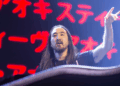 A DJ with long hair performs on stage, raising one hand in a rock gesture. Red Japanese characters are illuminated on a screen behind him. | FintechZoom