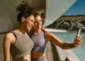 Two women in athletic wear smile and take a selfie together outdoors by a modern building and a pool on a sunny day. One has curly hair and an arm tattoo, while the other has earbuds. | FintechZoom