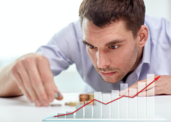 A man intently stacks coins on a table, with a rising bar graph and red upward trend line superimposed in the foreground, symbolizing financial growth or investment success. | FintechZoom