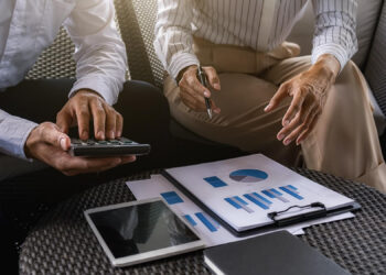 Two people sitting at a table review charts and graphs on paper. One uses a calculator, the other gestures while holding a pen. A tablet and clipboard are also on the table. | FintechZoom