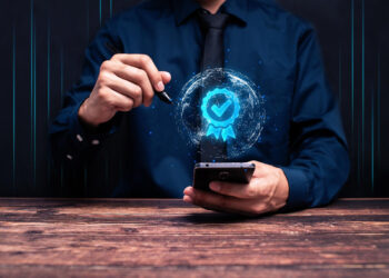 A person in a dark shirt and tie uses a smartphone with one hand and a stylus in the other. A glowing digital checkmark badge appears above the phone, symbolizing certification or digital approval. | FintechZoom