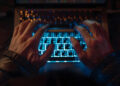 Close-up of two hands typing on a backlit laptop keyboard in a dimly lit environment, with blue light illuminating the keys and warm light reflecting off the hands. | FintechZoom