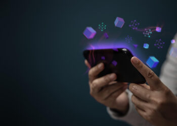 A person holding a smartphone with glowing digital cubes and network symbols emerging from the screen, representing technology or digital connections, against a dark background. | FintechZoom