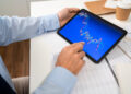 A person in a blue shirt uses a tablet displaying colorful financial or stock market charts, sitting at a desk with documents, a notebook, and a takeaway coffee cup. | FintechZoom