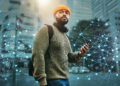 A man wearing a yellow beanie, green sweater, and headphones around his neck holds a smartphone while standing in a city. Digital network lines and nodes overlay the image, symbolizing connectivity. | FintechZoom