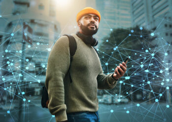 A man wearing a yellow beanie, green sweater, and headphones around his neck holds a smartphone while standing in a city. Digital network lines and nodes overlay the image, symbolizing connectivity. | FintechZoom