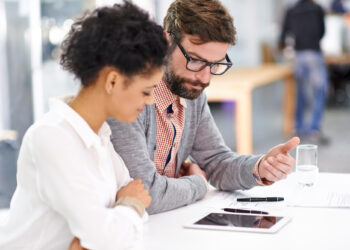 Two people sit at a desk looking at a tablet and documents. The man points at something on the tablet, and the woman sits beside him, both focused on the screen. A glass of water and papers are on the table. | FintechZoom