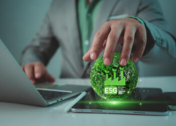 A person in a suit touches a glowing green holographic globe with "ESG" and sustainability icons, placed above a tablet on a desk next to an open laptop. | FintechZoom