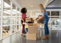Two women standing in a modern office, unpacking items from cardboard boxes on a table. One woman is wearing a pink top and pants, while the other wears a blue top and jeans. Large windows surround the bright space. | FintechZoom