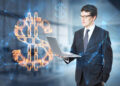 A man in a business suit holds a laptop while standing in an office environment. He looks at a large glowing digital dollar sign surrounded by network lines and symbols, representing finance and technology. | FintechZoom