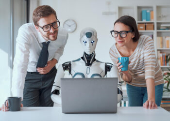 A man and a woman wearing glasses stand beside a humanoid robot at a desk, all looking at a laptop screen; the woman holds a blue mug. They appear engaged with what’s on the computer. | FintechZoom