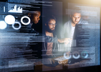 Three men collaborate at a computer surrounded by transparent digital screens displaying charts, graphs, and lines of code, suggesting teamwork and data analysis in a modern tech environment. | FintechZoom