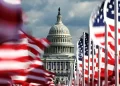 The U.S. Capitol building stands in the background with numerous American flags waving in the foreground on a cloudy day. | FintechZoom