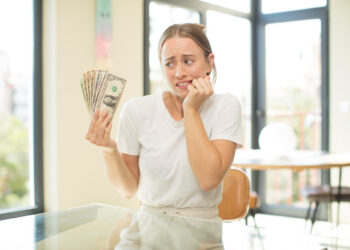 A worried woman in a white shirt sits at a table holding a fan of U.S. dollar bills, looking anxious and uncertain in a bright room with large windows. | FintechZoom