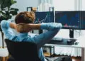 A person in a blue shirt sits at a desk with hands behind their head, looking at two computer monitors displaying stock market charts and financial data in a bright, modern office. | FintechZoom