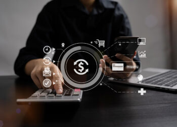 A person uses a calculator and holds a smartphone at a desk. Digital icons of money, shopping cart, email, and health are overlaid, representing online banking or digital financial transactions. | FintechZoom