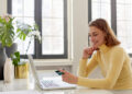 A woman in a yellow turtleneck sits at a desk with a laptop, smiling while holding a credit card. There are potted plants and large windows in the background, giving the room a bright, airy feel. | FintechZoom