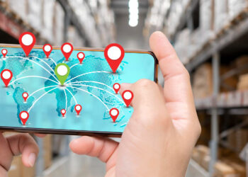 A person holds a smartphone in a warehouse, displaying a digital world map with multiple red location pins, suggesting global package tracking or logistics management. Shelves with boxes are blurred in the background. | FintechZoom