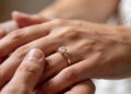 A close-up of one hand gently placing a gold ring with a prominent diamond on another person's ring finger, symbolizing an engagement or marriage. The background is softly blurred. | FintechZoom