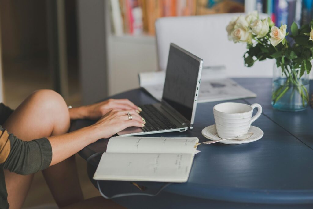 A person types on a laptop at a blue table with an open notebook, a cup of coffee on a saucer, and a vase of white roses. Shelves with books are visible in the blurred background. | FintechZoom