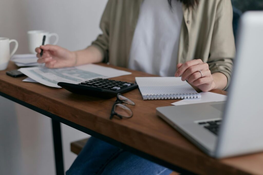 A person sits at a desk with a laptop, calculator, notebook, glasses, and documents. They are writing with a pen and reviewing paperwork, appearing to work or study. Two mugs are also on the desk. | FintechZoom