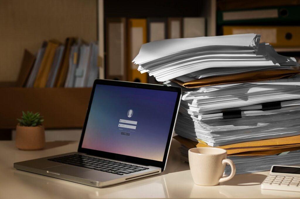 A laptop with a login screen sits on a desk beside a large stack of papers, a coffee mug, a small plant, and a keyboard, with shelves of folders in the background. | FintechZoom