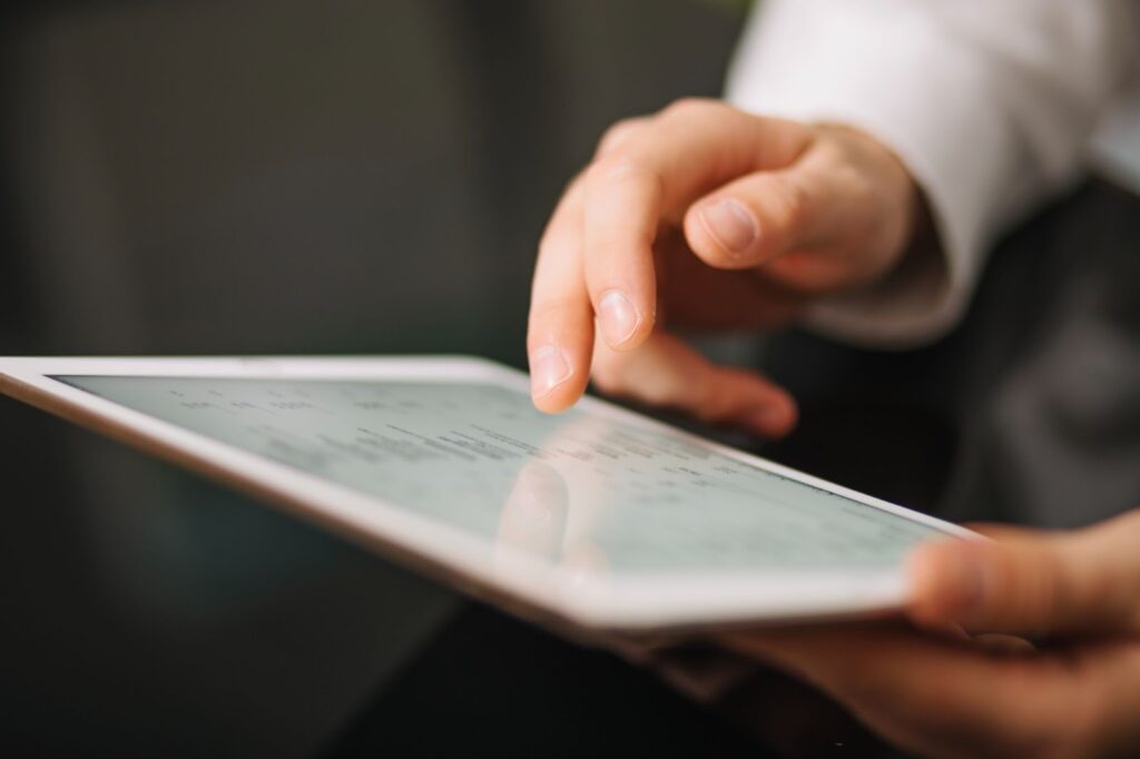 A person uses their index finger to navigate a touchscreen tablet, which displays a bright white screen with indistinct text or graphics. The background is blurred, focusing attention on the hand and device. | FintechZoom