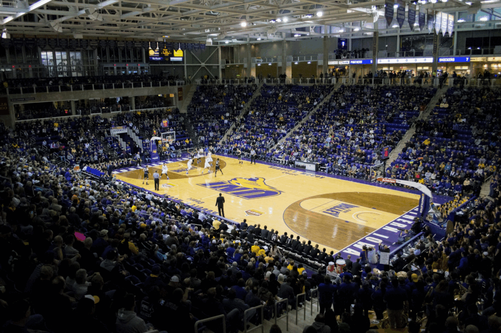 A wide view of a packed indoor basketball arena, with players on the court during a game. The stands are filled with spectators, and bright lights illuminate the wooden floor and surrounding seating areas. | FintechZoom