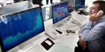 A man in a business shirt talks on a phone while looking at large computer monitors displaying financial graphs and stock market data in an office setting. | FintechZoom