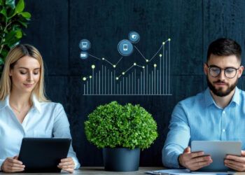 Two people sit at a table using digital tablets, with a potted plant between them. A digital chart with data points and technology icons is displayed in the background, suggesting data analysis or business growth. | FintechZoom