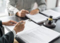 Two people sit at a table reviewing legal documents, with one hand holding a pen and the other gesturing. A judge’s gavel and case files are visible on the desk. | FintechZoom