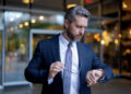A man in a suit stands outside a building, holding his glasses in one hand and looking at his watch. He appears focused and professional, with short grey hair and a trimmed beard. | FintechZoom