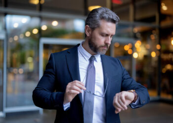 A man in a suit stands outside a building, holding his glasses in one hand and looking at his watch. He appears focused and professional, with short grey hair and a trimmed beard. | FintechZoom