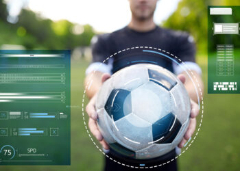 A person holds a football towards the camera on a grassy field, with digital graphics and data overlays suggesting sports analysis or futuristic technology. The background is blurred and outdoors. | FintechZoom