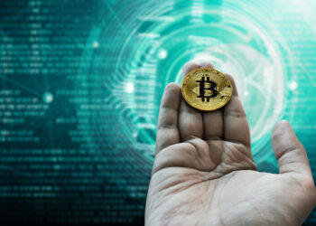 A hand holds a gold-coloured Bitcoin coin against a digital background with blue-green tones and abstract circular tech patterns, symbolising cryptocurrency and blockchain technology. | FintechZoom