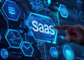 A hand points towards a digital screen displaying the word "SaaS" among various technology-related blue icons, representing cloud computing and software as a service concepts. | FintechZoom