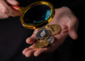 A person holds several cryptocurrency coins, including Ethereum, in one hand while examining them with a magnifying glass in the other hand. The background is dark. | FintechZoom