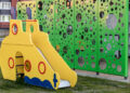 A yellow playground slide shaped like a submarine sits on grass in front of a green fence with variously sized circular cut-outs. | FintechZoom