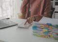 A person in a pink blazer uses a calculator at a desk with a laptop, a stack of documents organised with colourful tabs, and a cup in the background. | FintechZoom