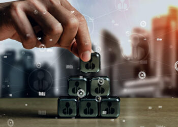 A hand stacks transparent cubes with human icons into a pyramid shape on a table, with a blurred cityscape and digital network graphics in the background, symbolising team building and business networking. | FintechZoom