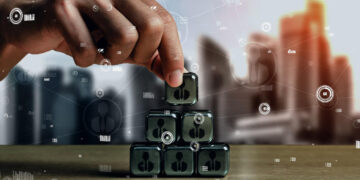 A hand stacks transparent cubes with human icons into a pyramid shape on a table, with a blurred cityscape and digital network graphics in the background, symbolising team building and business networking. | FintechZoom